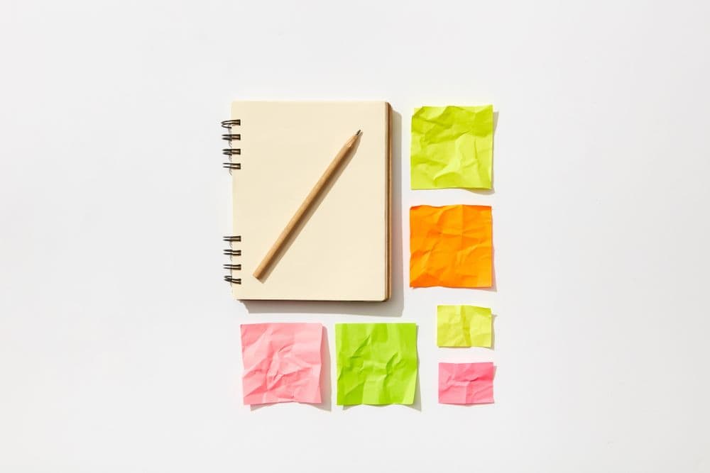 A minimalist flat lay featuring a spiral notepad, pencil, and colorful crumpled sticky notes on a white surface.